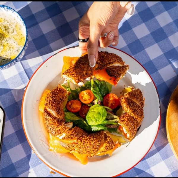 Hand reaching for a Turkish simit sandwich with cheese, greens, and tomatoes on a blue checkered tablecloth.