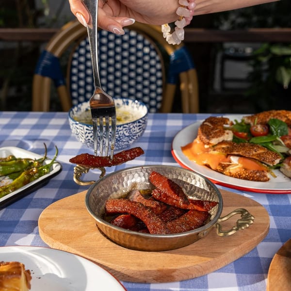 A hand uses a fork to pick up a piece of Turkish sucuk from a copper pan on a checkered tablecloth.