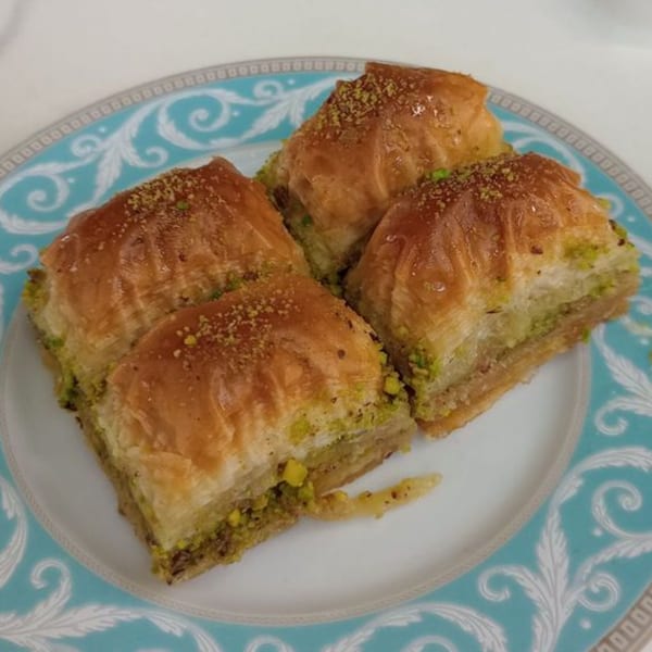 Four pieces of golden-brown baklava, sprinkled with green pistachios, on a decorative blue and white plate.