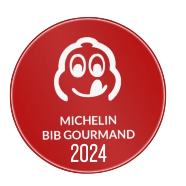 Red circular Michelin Bib Gourmand 2024 award logo with a white smiling face.