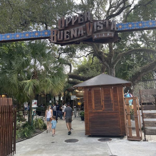 A large sign for "Upper Buena Vista Miami" hangs over a pathway with people and lush greenery.