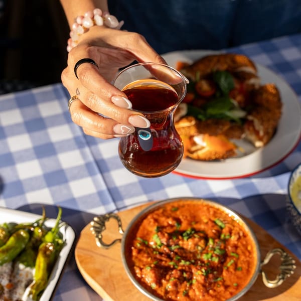 A hand holds a Turkish tea glass with an evil eye, over a table with Turkish dishes.