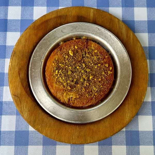 Turkish kunefe dessert, a golden pastry topped with pistachios, served on a metal plate over a blue and white checkered tablecloth.