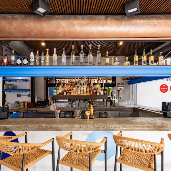 A modern bar with a concrete counter, wicker stools, and shelves of liquor bottles.