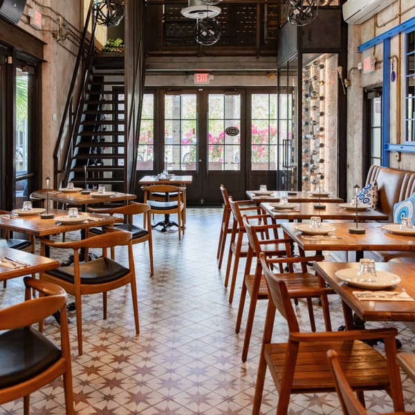 A restaurant interior with wooden tables and chairs, a staircase, and large windows with flowering plants outside.