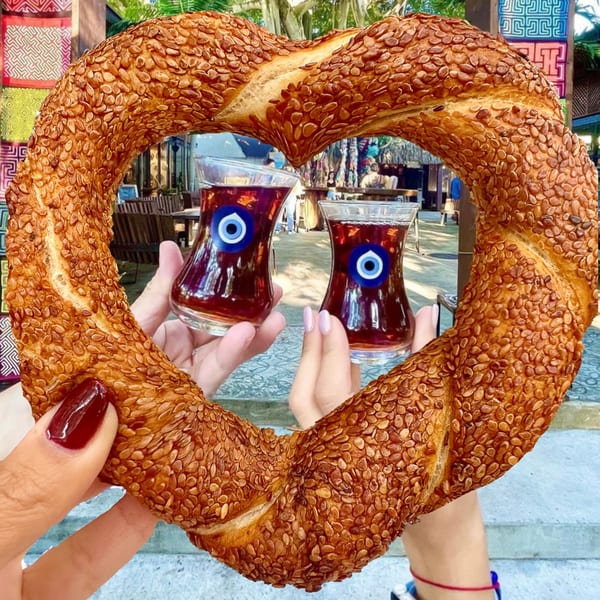 Heart-shaped Turkish simit with sesame seeds, framing two hands holding Turkish tea glasses with evil eye charms.